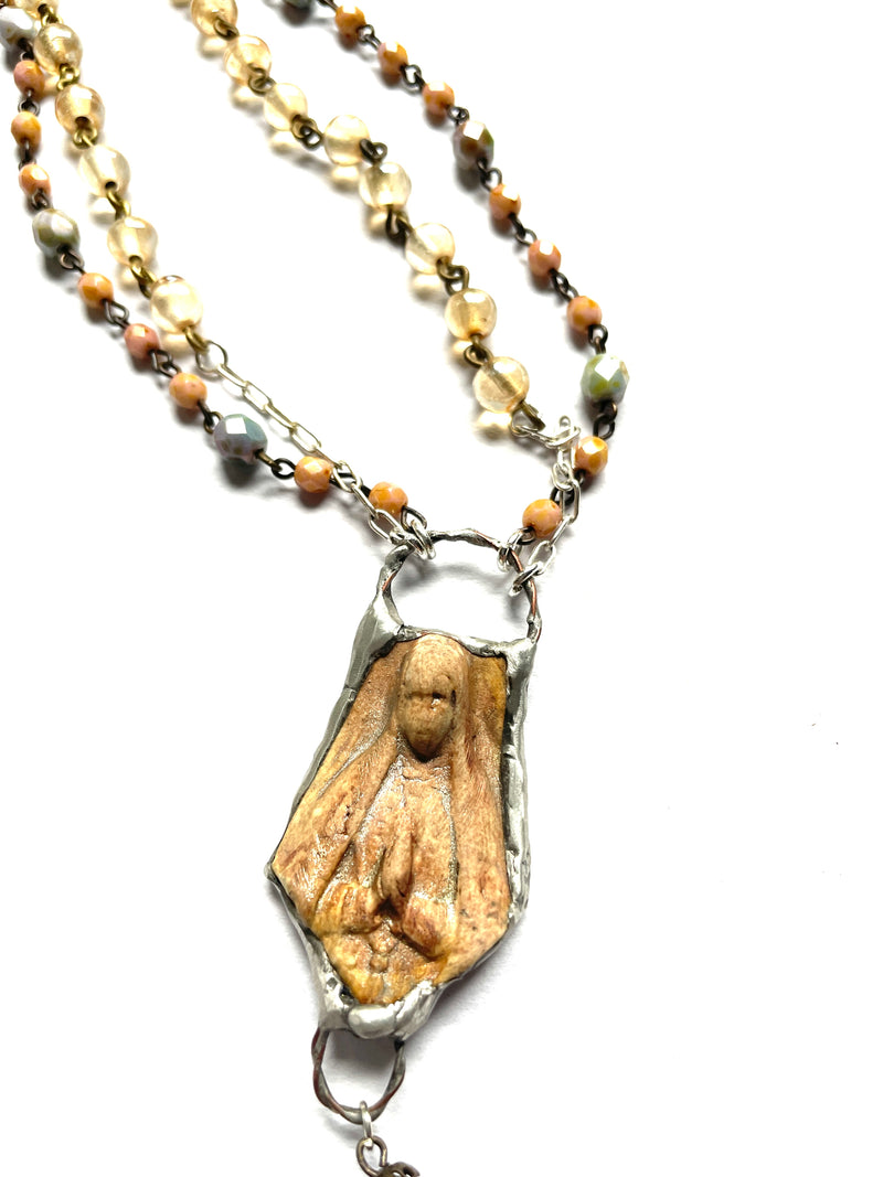 close up of 2 layers of beaded chain necklace. peach, green, gold glass beads, a single pearl and a pewter silver bezel wraps a smokey quartz crystal point. silver chain, clasp and beaded extender, ceramic pendant of the Virgin Mary, golden brown in color, in a pewter bezel